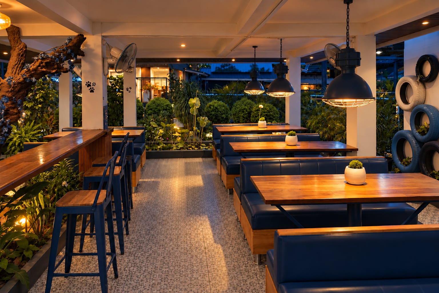 The café in the evening: long wooden tables and blue benches under black pendant lamps, open to the lit garden