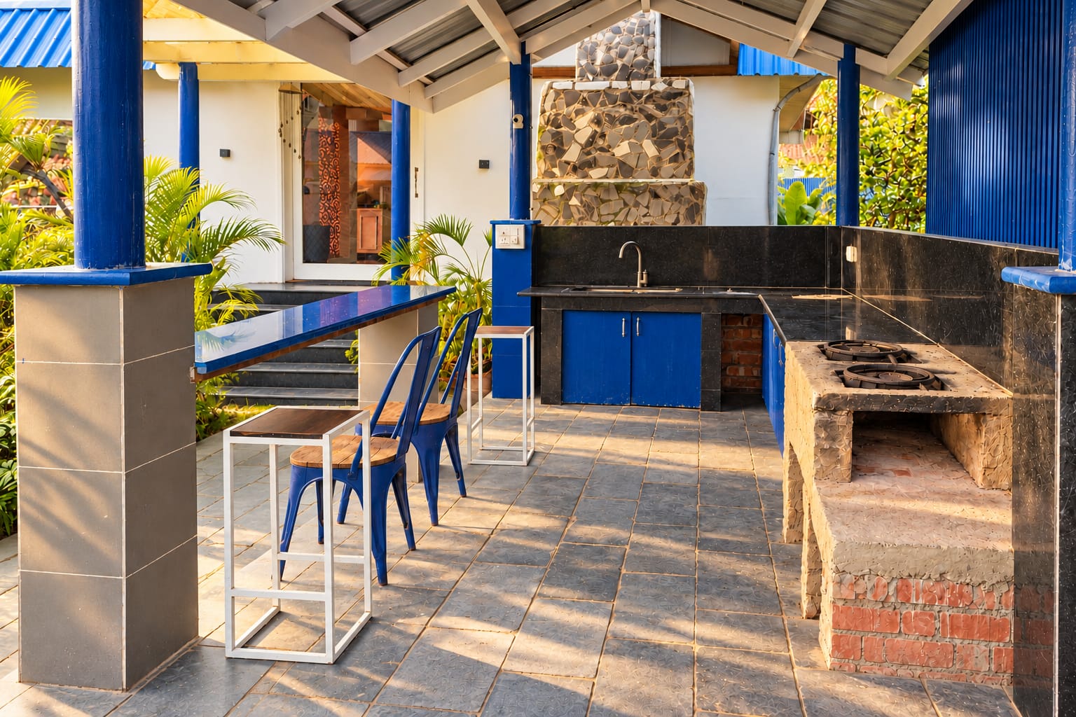 The outdoor counter under a blue-roofed shelter: a tiled worktop with a brick grill and blue bar stools along it